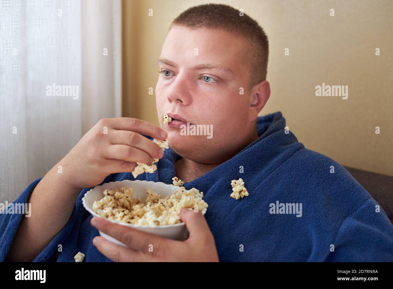 Fat man in a robe eating popcorn while sitting on the couch, obesity