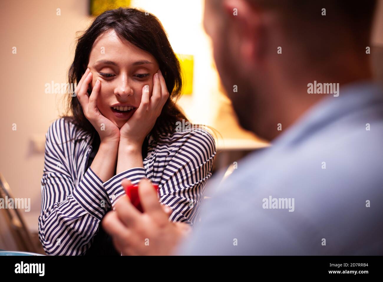 Woman looking happy at engagement ring during marriage proposal while having romantic dinner. Man asking his girlfriend to marry in the kitchen during romantic dinner. Happy caucasian woman smiling being speechless Stock Photo