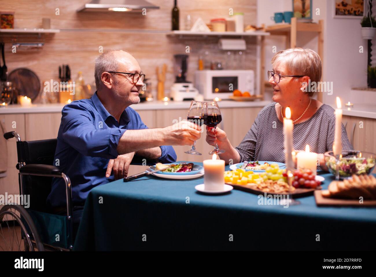 Handicapped man having dinner with man in kitchen toasting. Wheelchair immobilized paralyzed handicapped man dining with wife at home, enjoying the meal Stock Photo