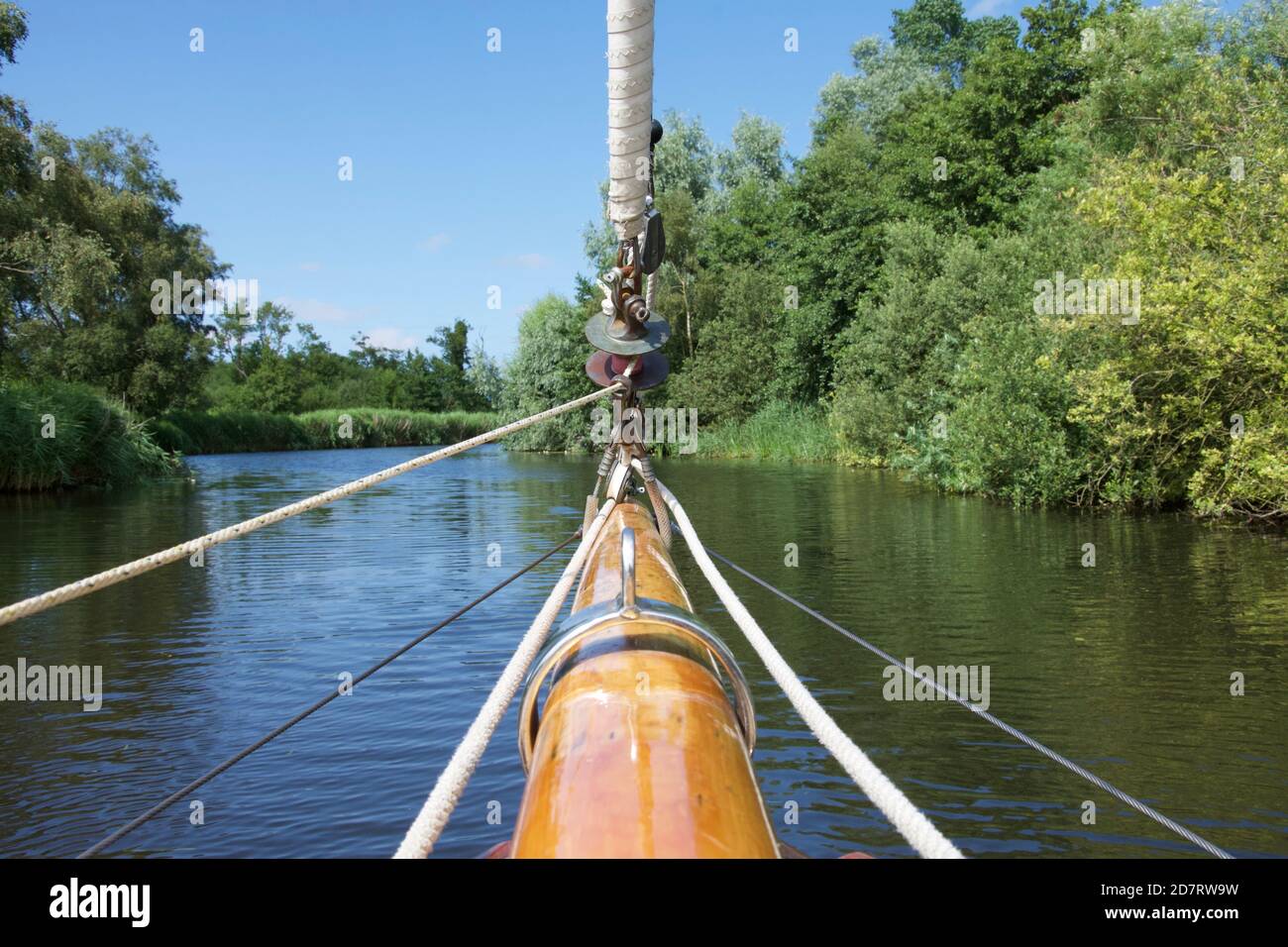 The wooden bowsprit and furled jib of a sailing yacht, moving up the centre of a sunny river between tree-lined banks Stock Photo