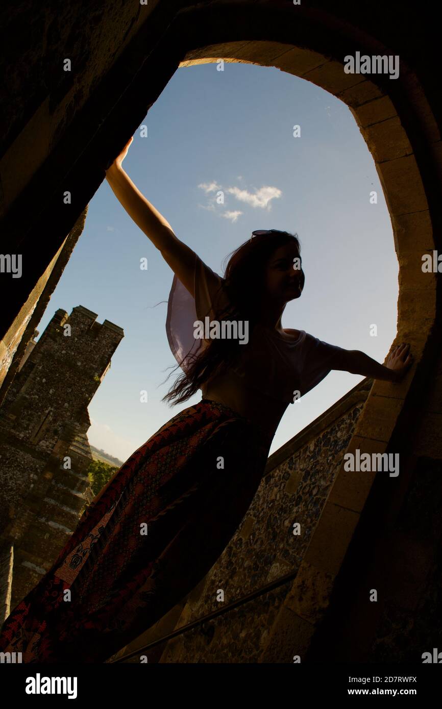 Silhouette of a thin young lady with arms outstretched, posing in the archway (open doorway) of a castle in the evening light. Stock Photo