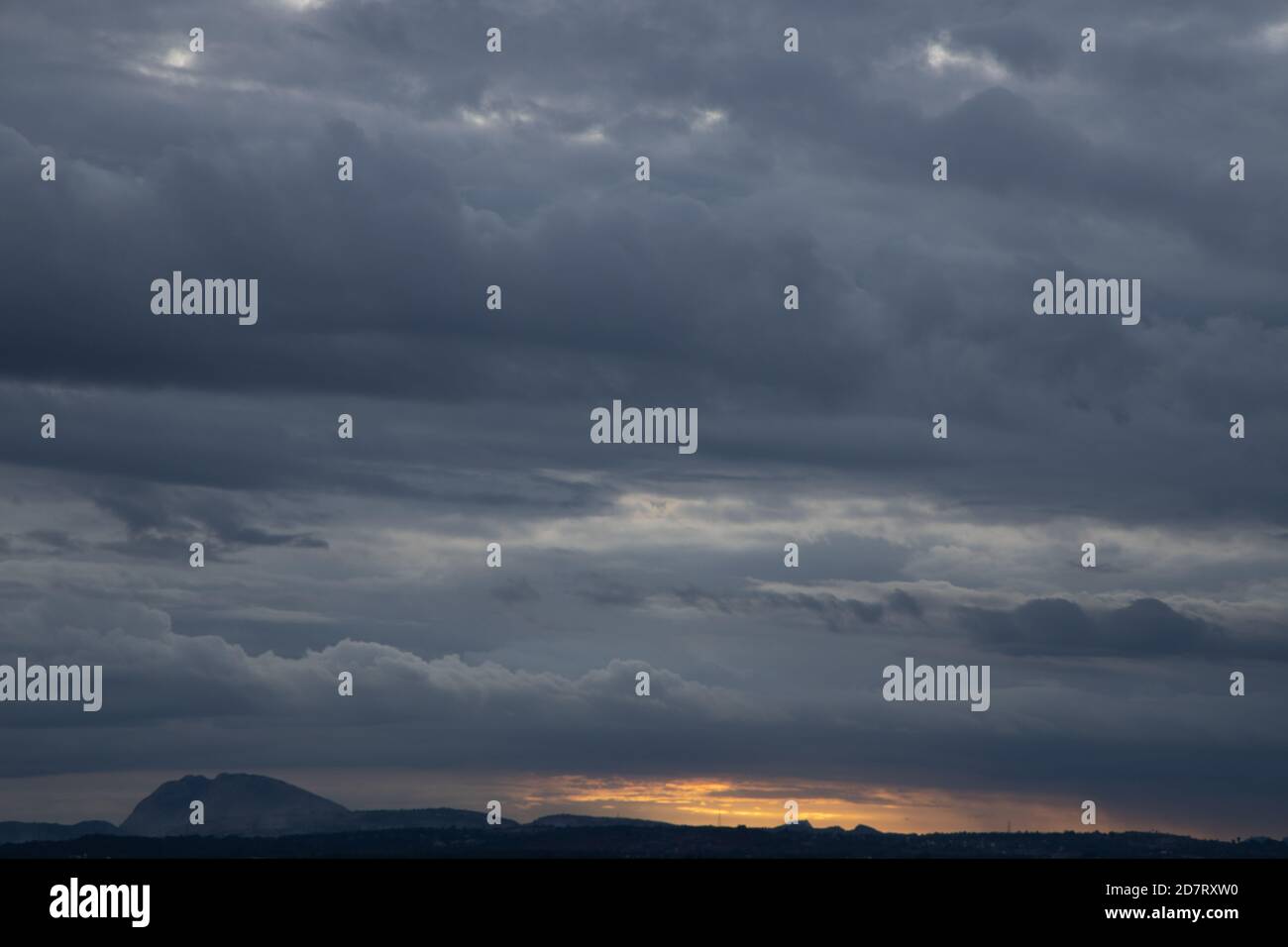 Dark rain clouds in the horizon with Silhouette of  hills in the bottom with golden light peeping in Stock Photo