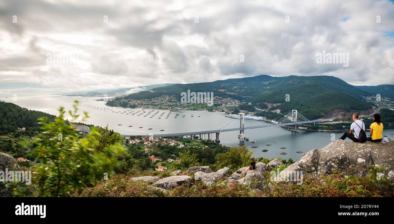 A non-recognisable couple enjoys the sunset over the Ria de Vigo estuary in Redondela on a cloudy Summer afternoon, with the recently extended Rande b Stock Photo