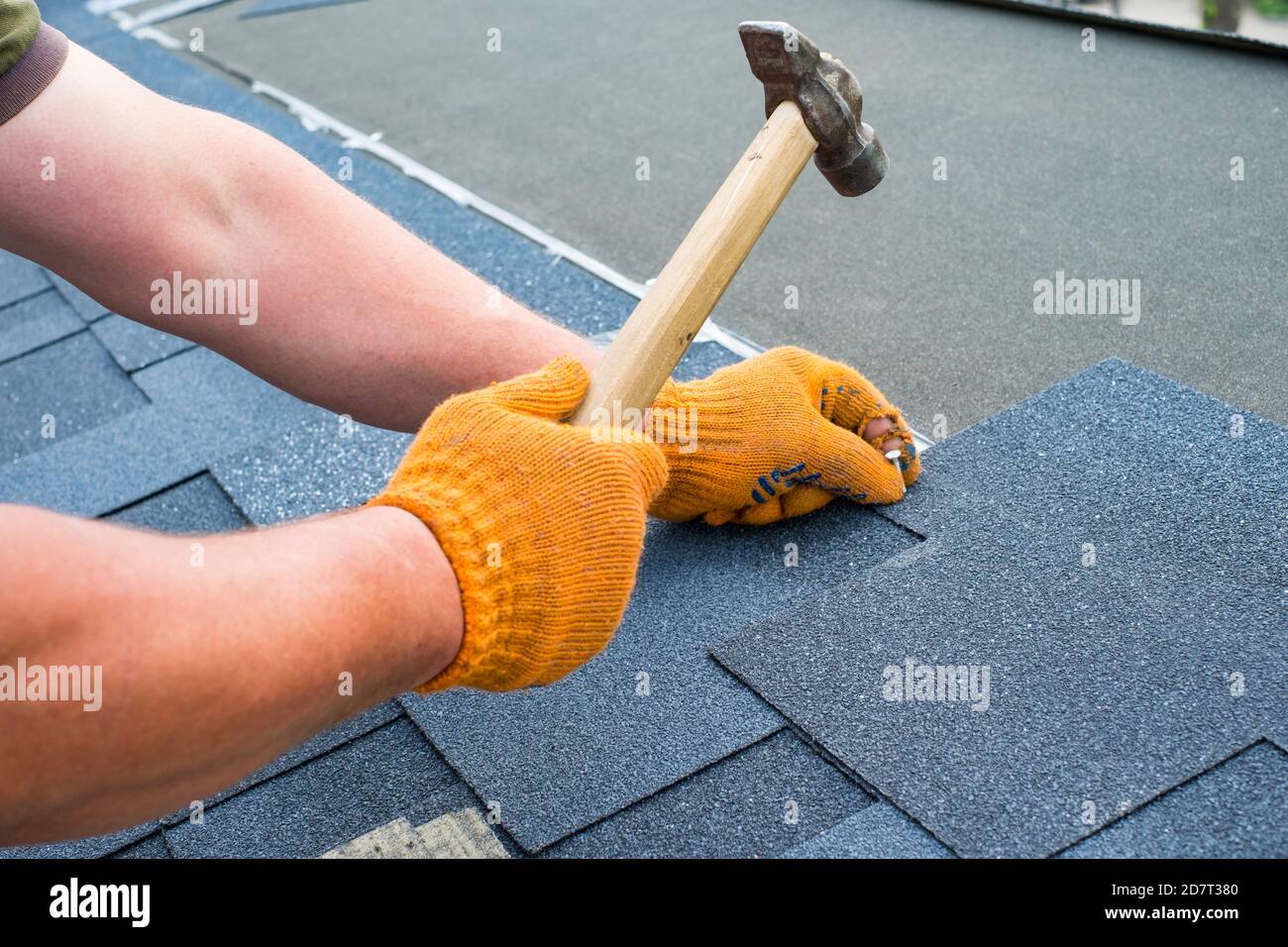 Workers hands installing bitumen roof shingles using hammer in nails