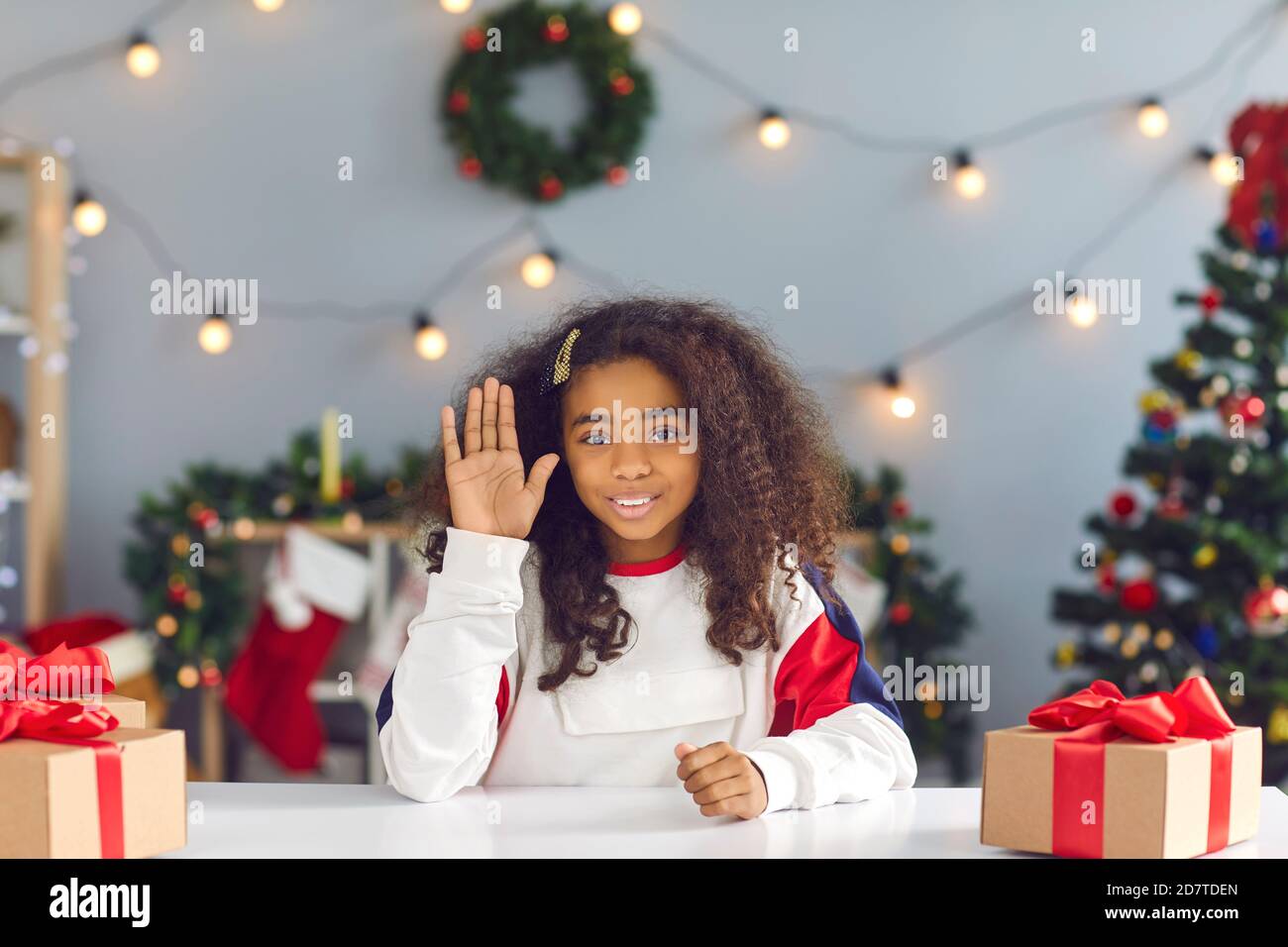 Cute african american girl sits at a table with gifts in front of a webcam and waves a hand. Stock Photo