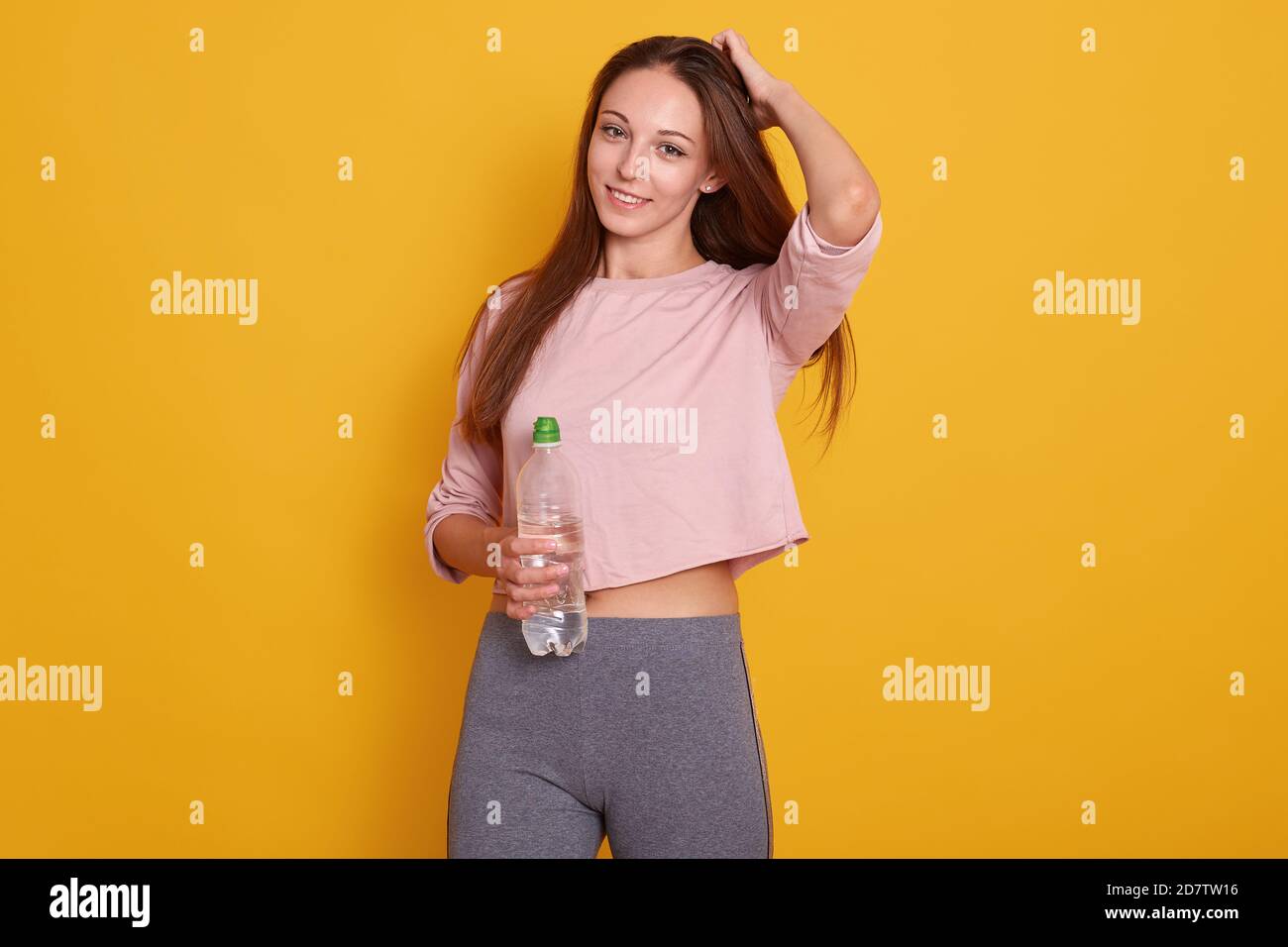 Studio shot of caucasian fitness woman model standing in sporty gym clothing, having dark straight hair, holds water bottle, posing isolated over yell Stock Photo