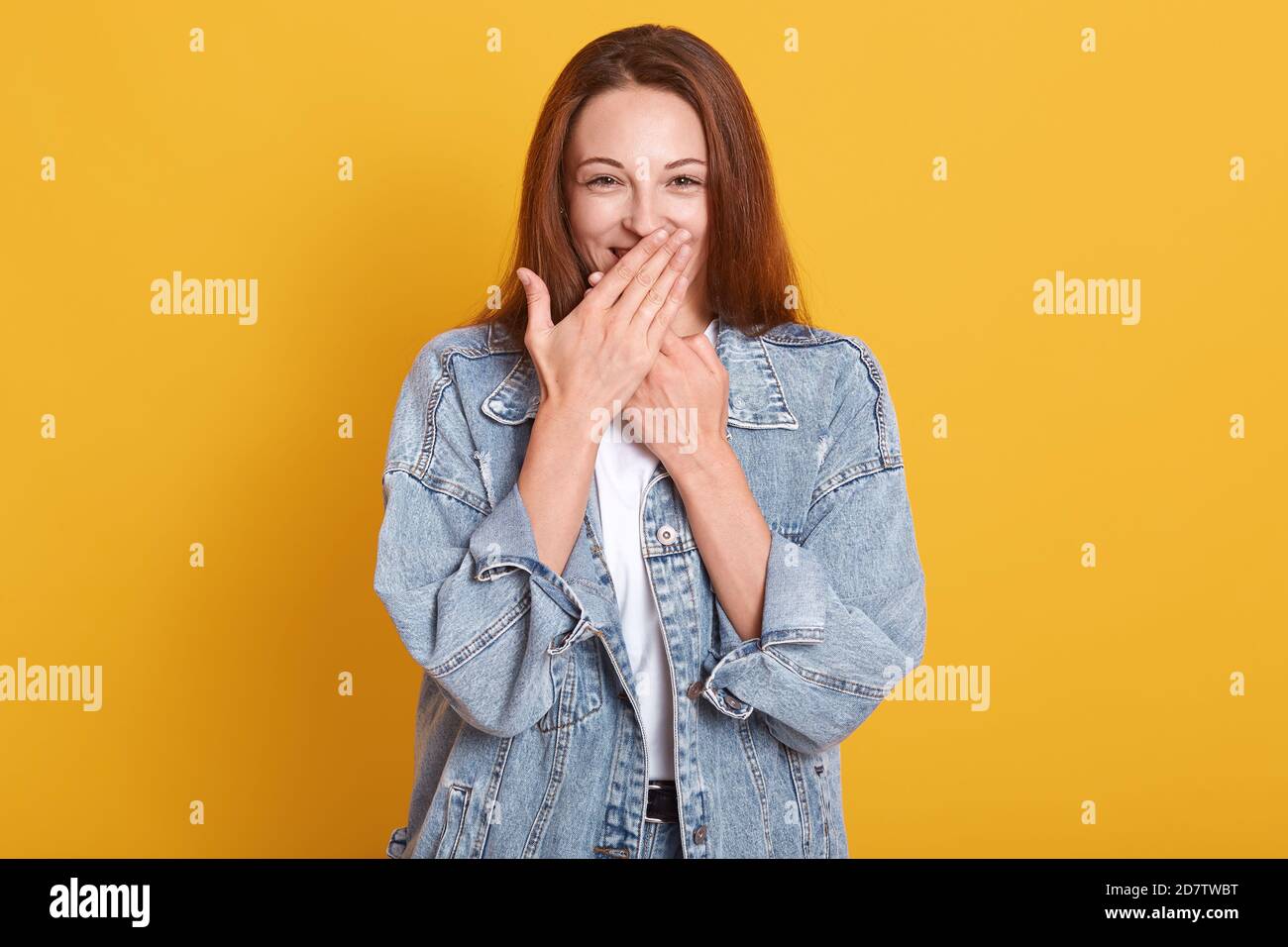 Happy good looking young model covering her mouth with both hands, having shy surprised look, being in high spirits, wearing denim jacket and white t Stock Photo