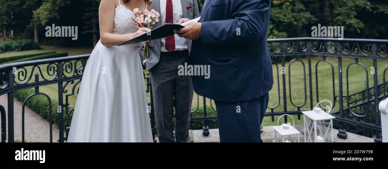 Bridal couple signs official marriage cerificate in hands of a registrar. Young bride wears white dress and holds wedding bouquet with pink roses. Cla Stock Photo