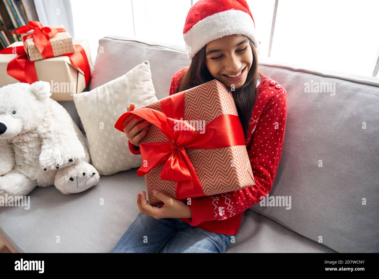 Happy cute indian kid girl holding gift box Christmas present sitting on couch. Stock Photo