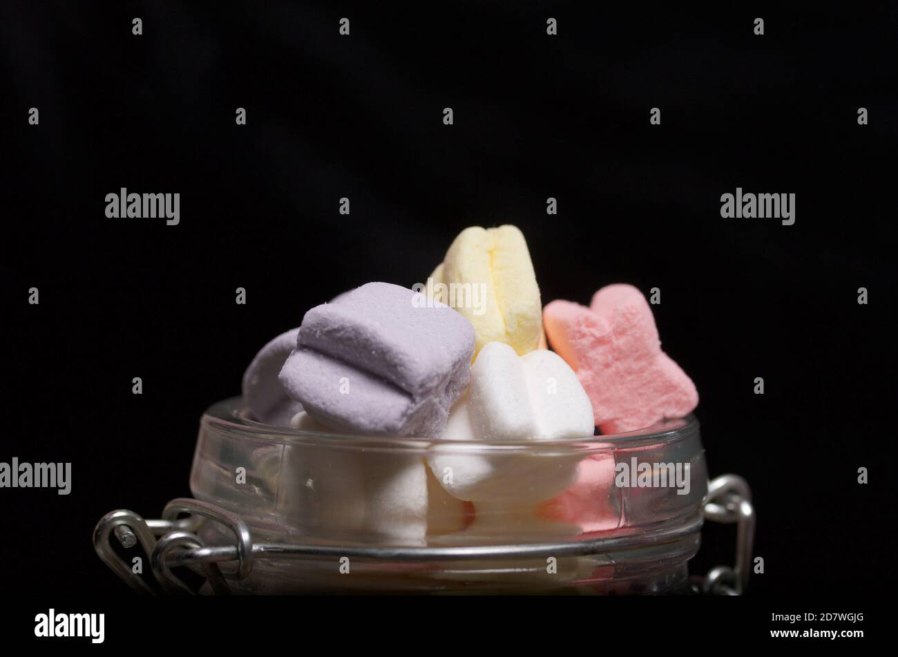 Marshmallows of different colors. They lie in glass jar. Photographed close-up on a black background. Stock Photo