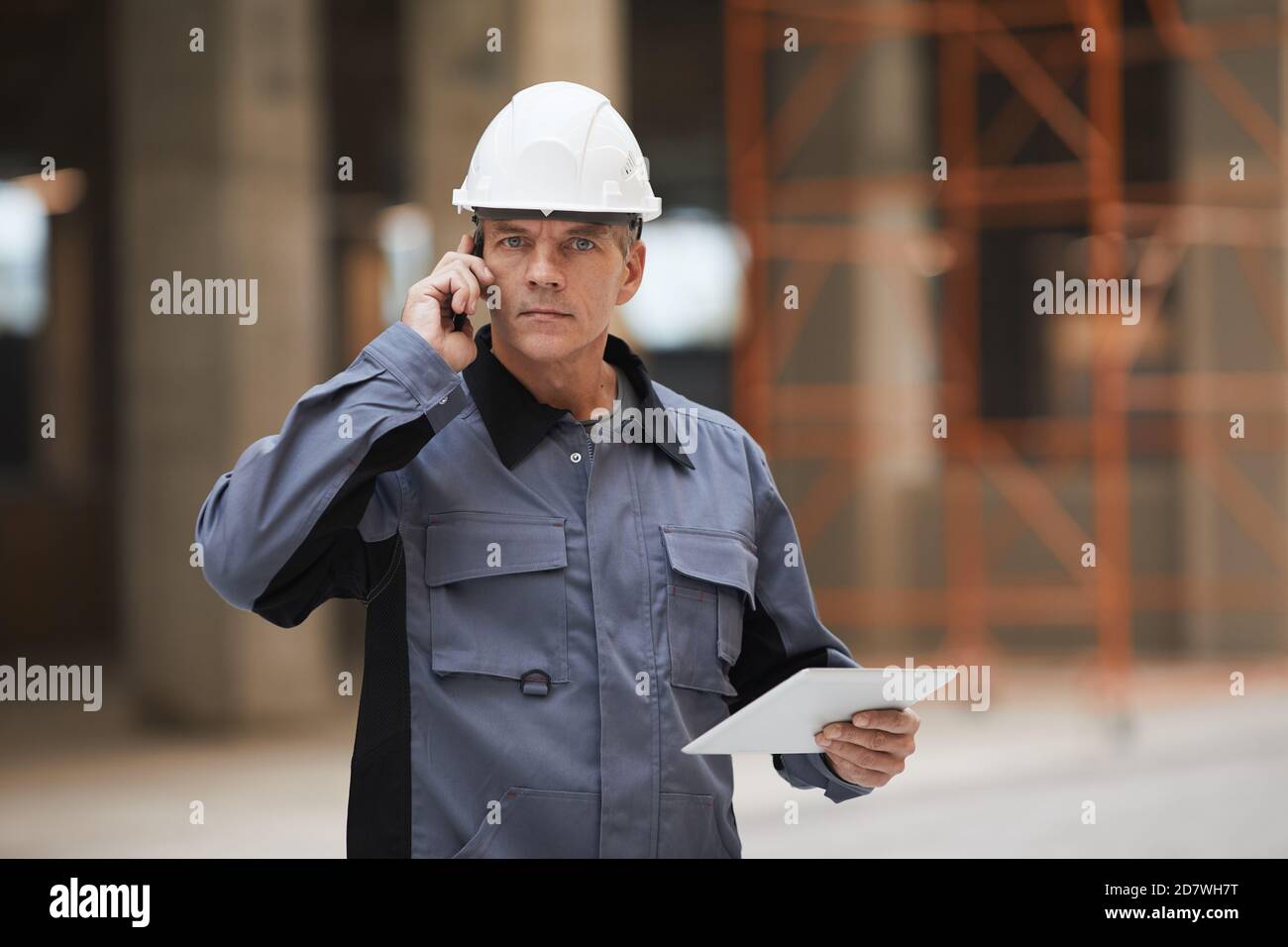 Waist up portrait of mature worker speaking by smartphone and looking at camera while standing at construction site or in industrial workshop, copy space Stock Photo