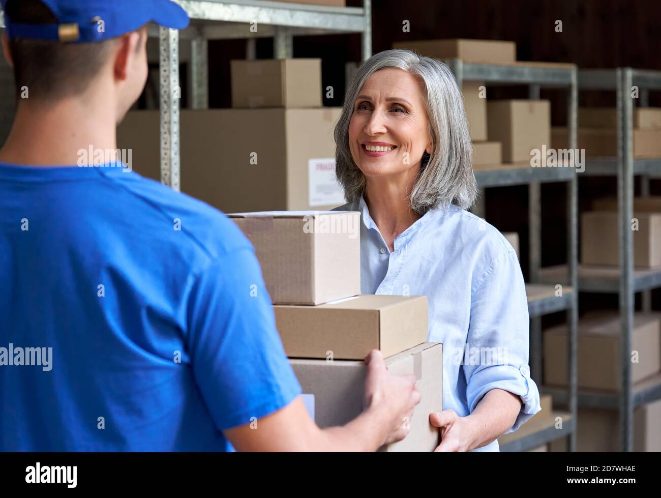 Happy mature female manager giving parcels boxes to courier in warehouse. Stock Photo
