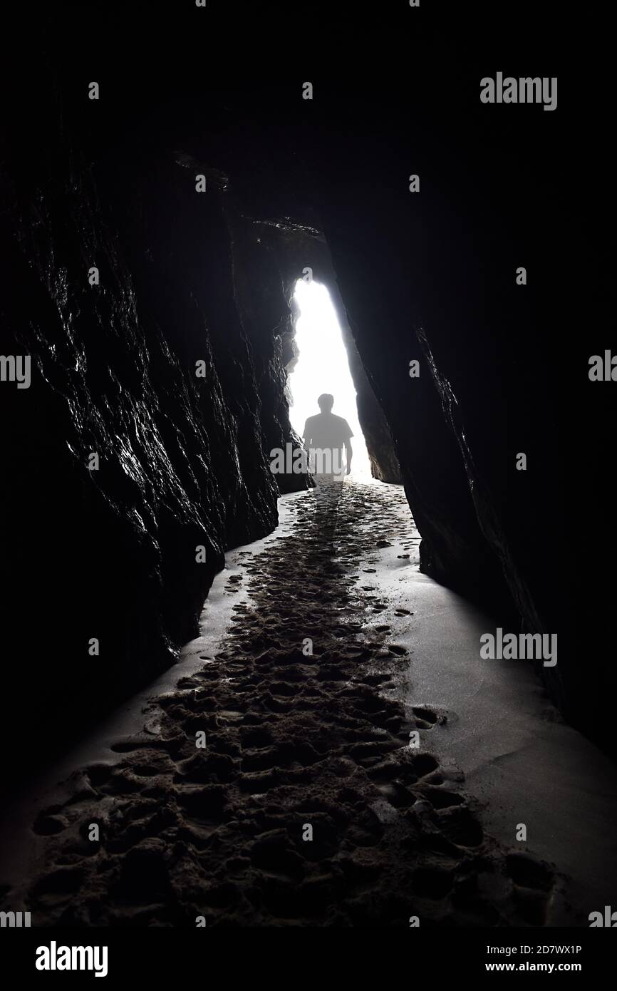 A transparent ghostly figure walks towards the light at the end of a tunnel, heading out of a cave with sandy footprints leading out from the front. Stock Photo