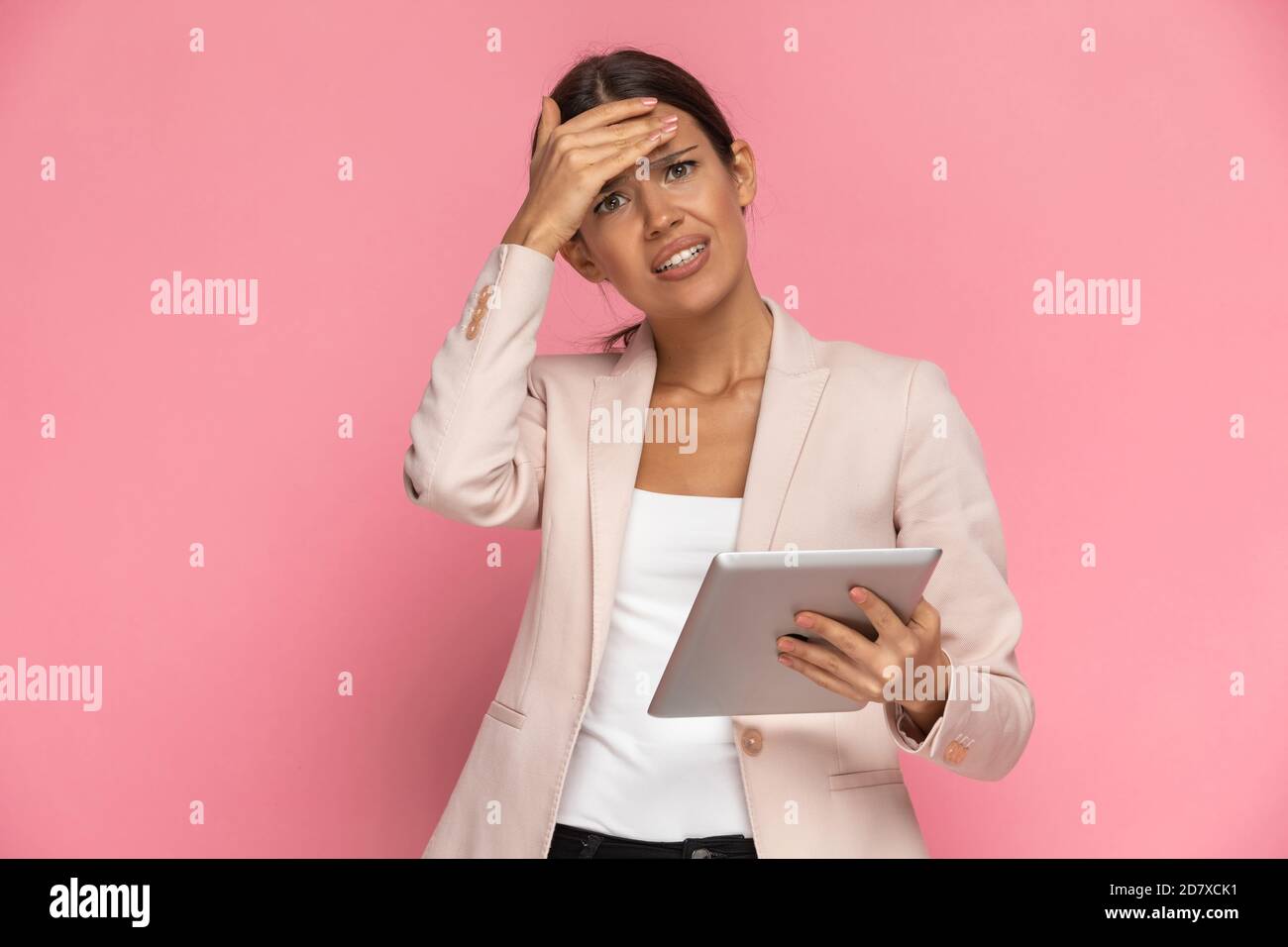 beautiful businesswoman holding her tablet and slapping her face, feeling anxious on pink background Stock Photo