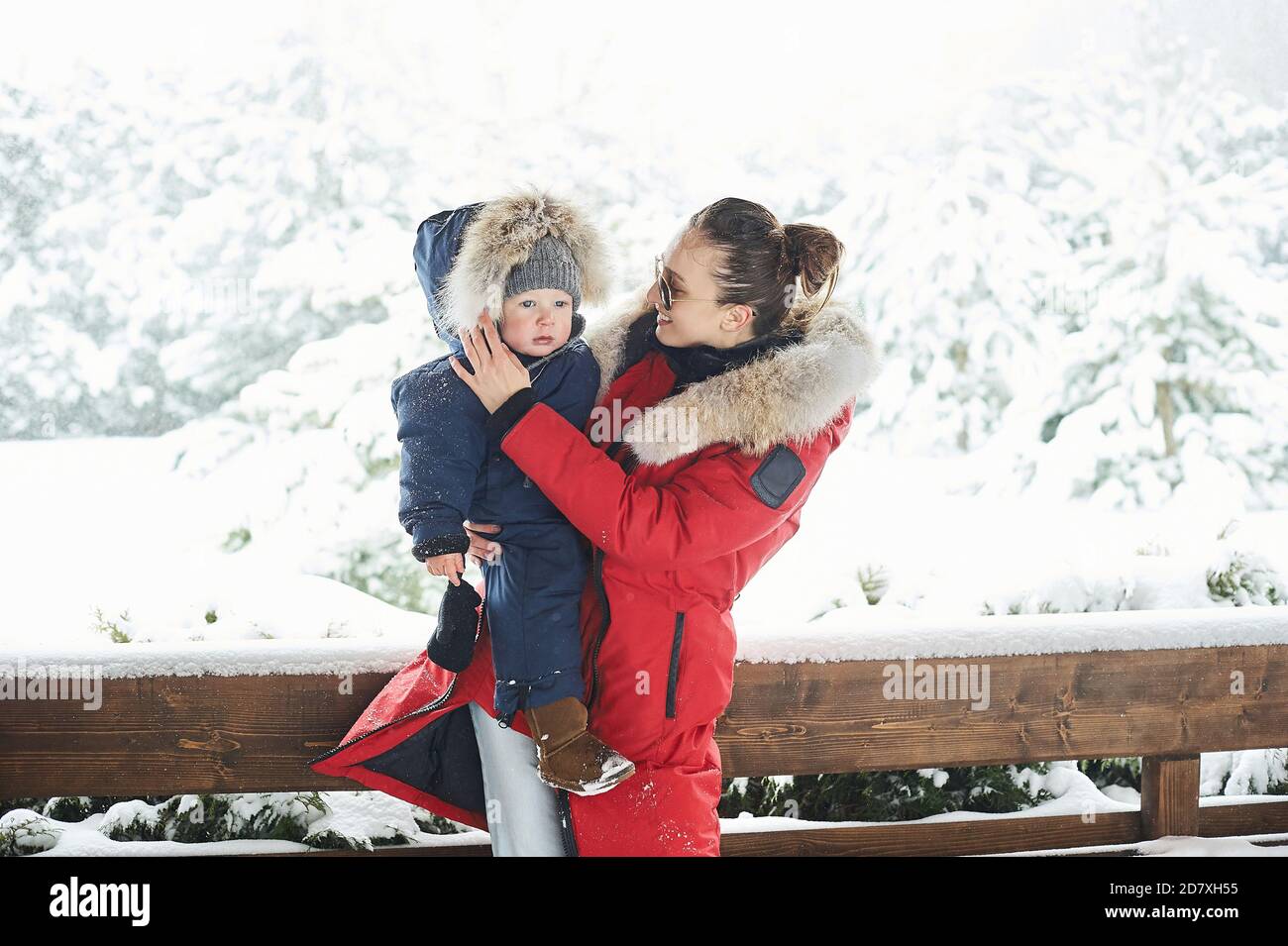 Beautiful young mother dressed in red jacket with a child in winter near the house with a snow covered christmas tree Stock Photo