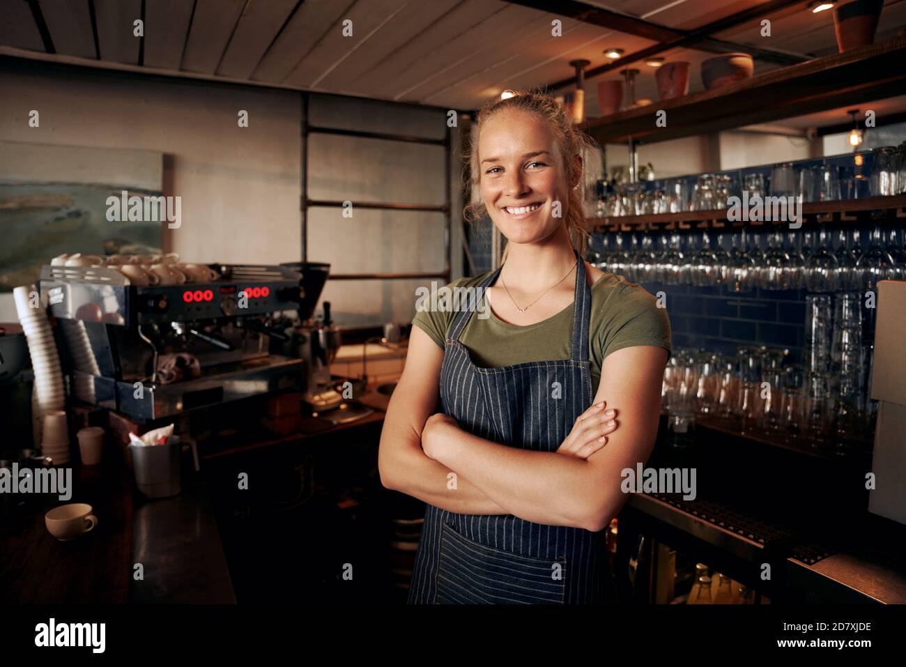 Portrait of young female waitress wearing apron standing behind counter in cafe with crossed hands Stock Photo