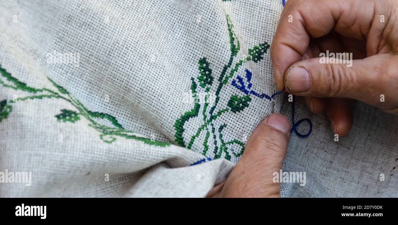 Hands of an elderly woman embroidering a cross-stitch floral pattern on linen fabric. Embroidery, handwork, needlecraft concept. Close-up Stock Photo