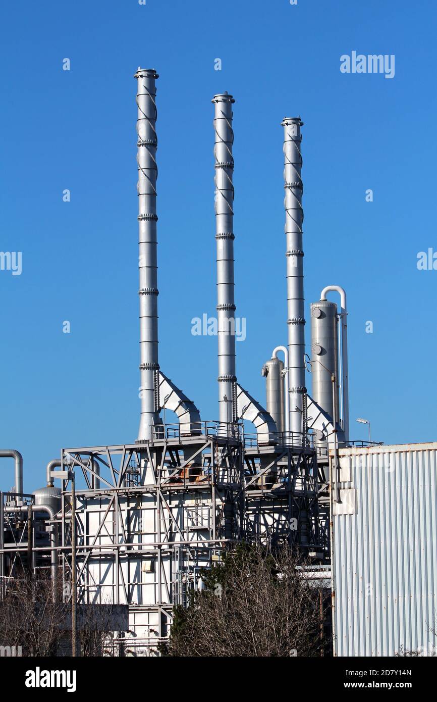 Three brand new shiny metal industrial chimneys rising high above modernized part of large oil refinery industrial complex surrounded with trees Stock Photo