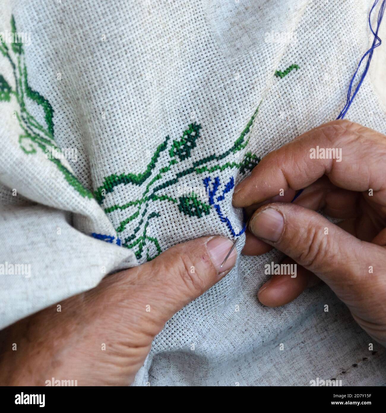 Hands of an elderly woman embroidering a cross-stitch floral pattern on linen fabric. Embroidery, handwork, needlecraft concept. Close-up Stock Photo