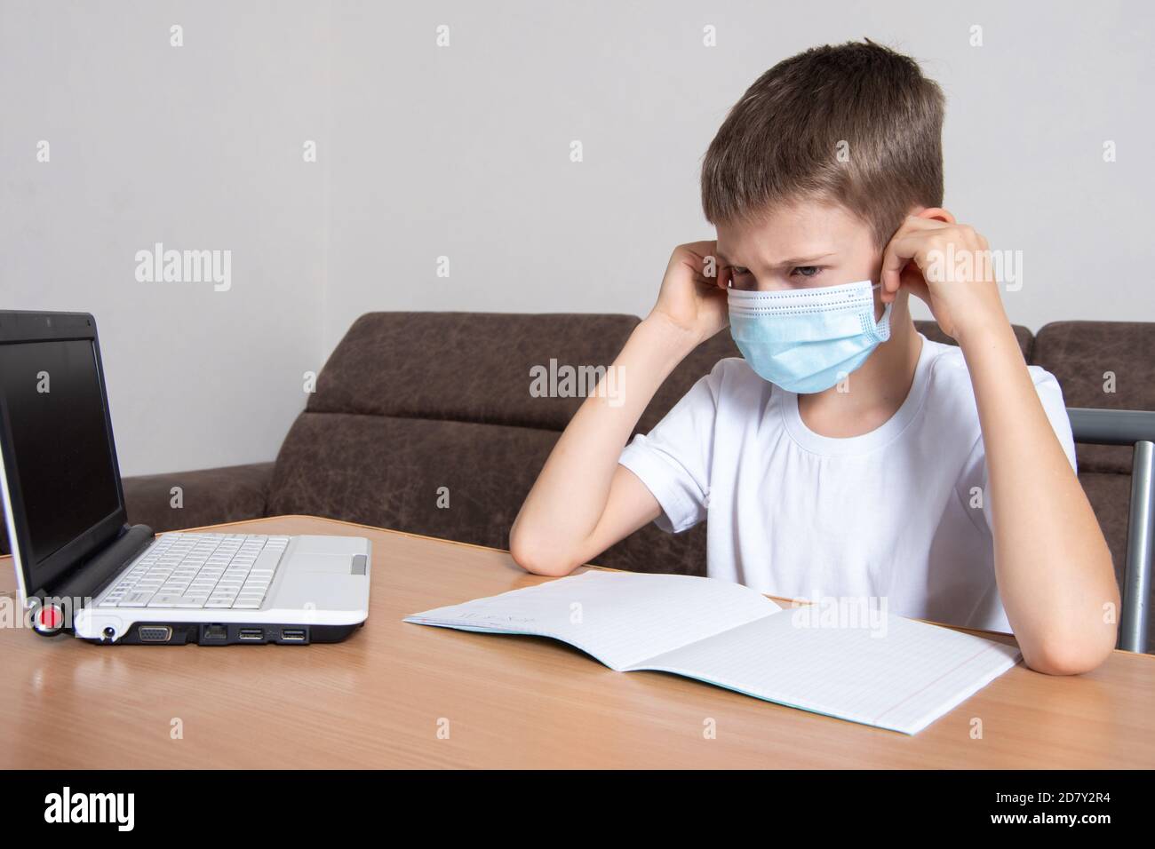 A child in a protective mask on his face looks displeasedly at a laptop, a boy learns remotely online at home Stock Photo