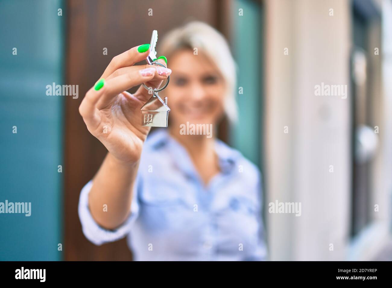 Young blonde woman smiling happy holding key of new house at the city ...