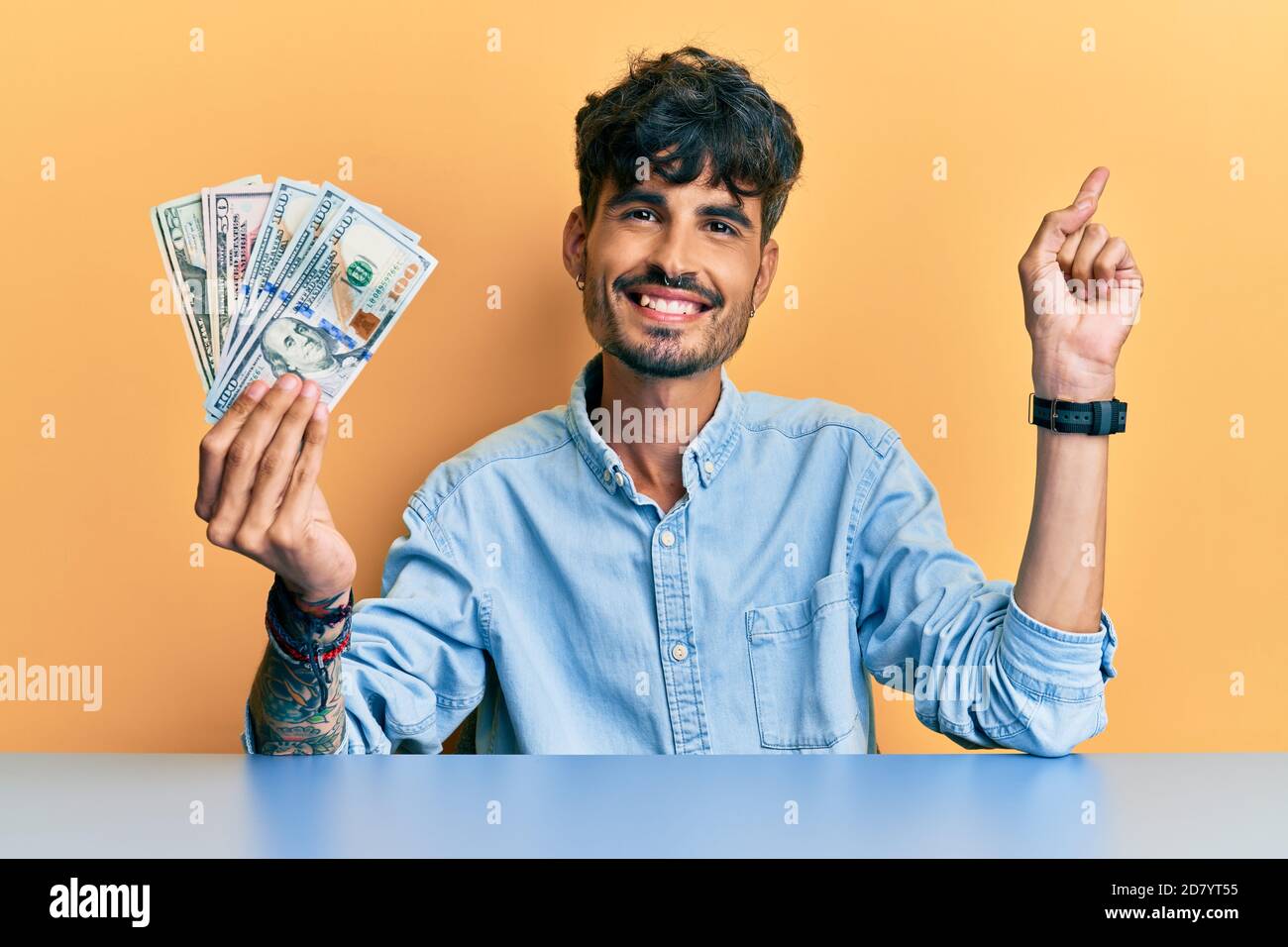 Young hispanic man holding dollars sitting on the table smiling happy pointing with hand and finger to the side Stock Photo