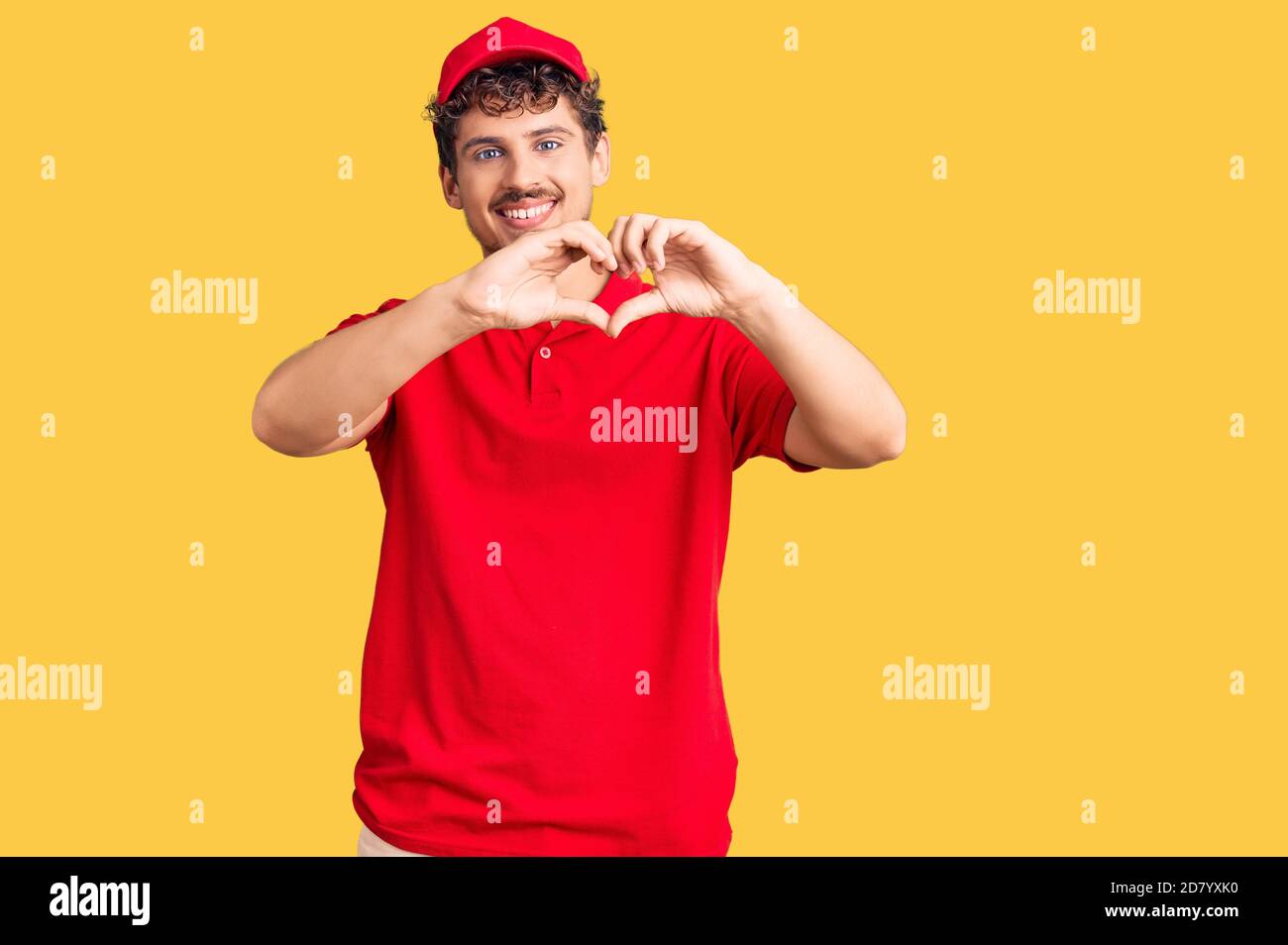 Young handsome man with curly hair wearing delivery uniform smiling in love showing heart symbol and shape with hands. romantic concept. Stock Photo