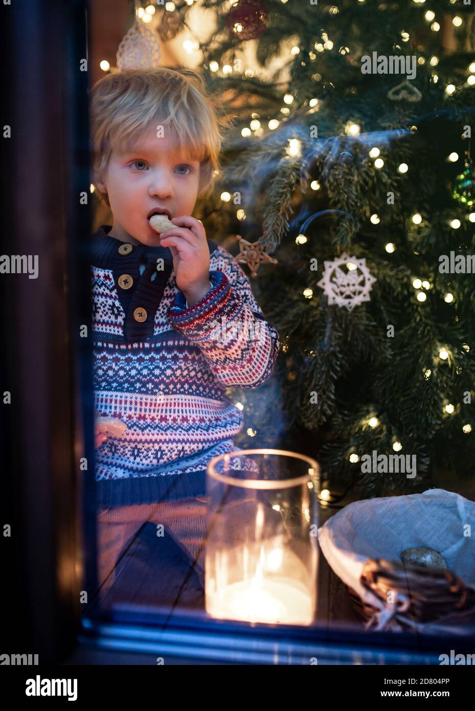 Portrait of small boy indoors at home at Christmas, eating by window at night. Stock Photo