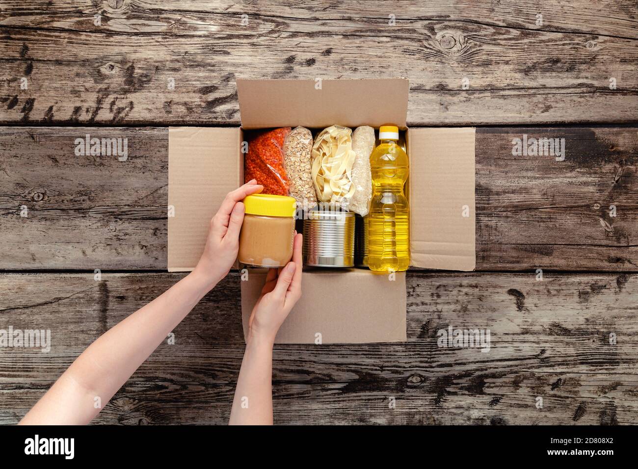 Person woman receiving donation Food box. Female volunteer hands packing donation box with food items of staple products on wooden table. Donate Food Stock Photo