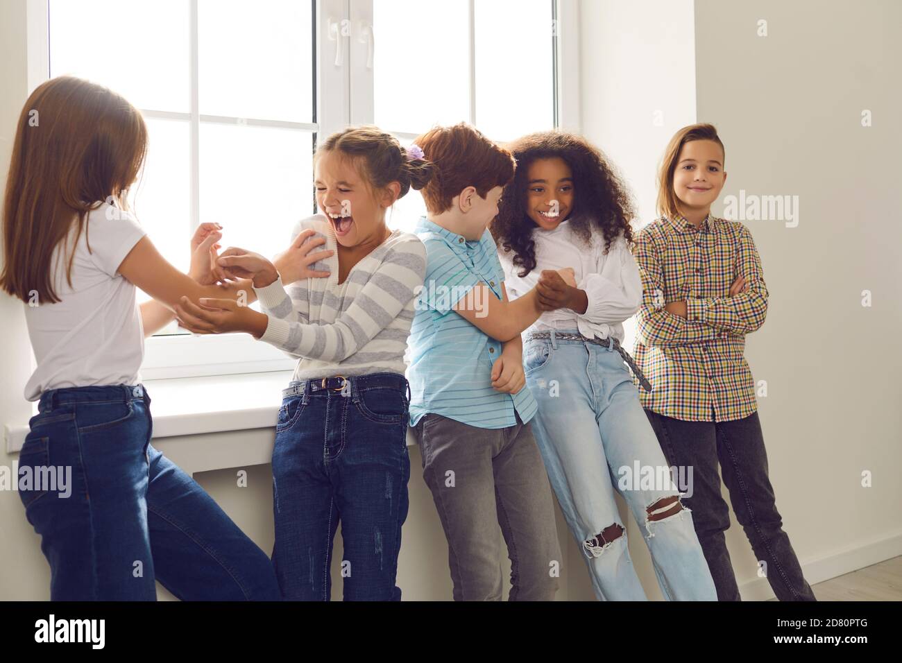 Active group of multiethnic children have fun together and tickle each other standing by the window. Stock Photo