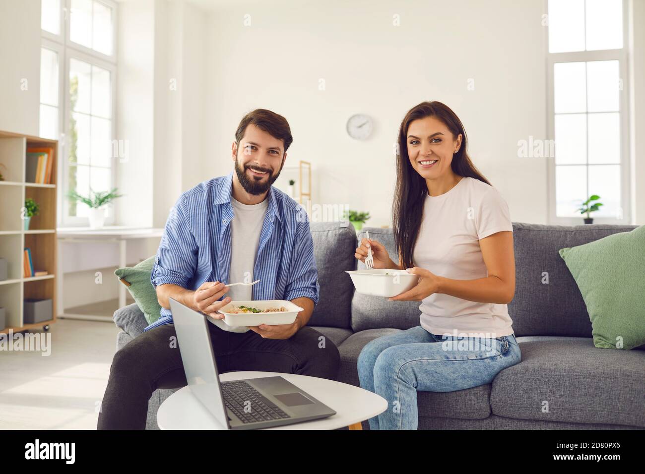 Man and woman holding takeaway food and having lunch at home sitting on sofa in front of laptop. Stock Photo