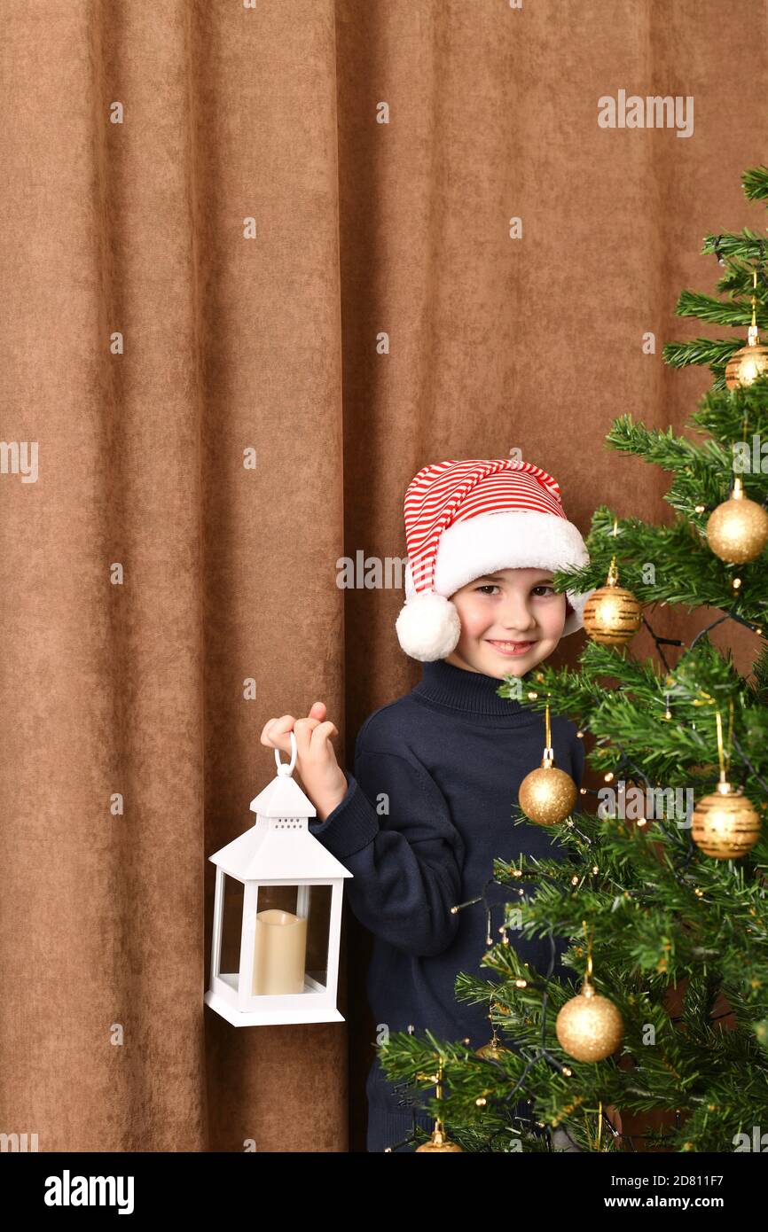 A child with a lantern in his hand stands embarrassedly smiling near a dressed Christmas tree. Stock Photo