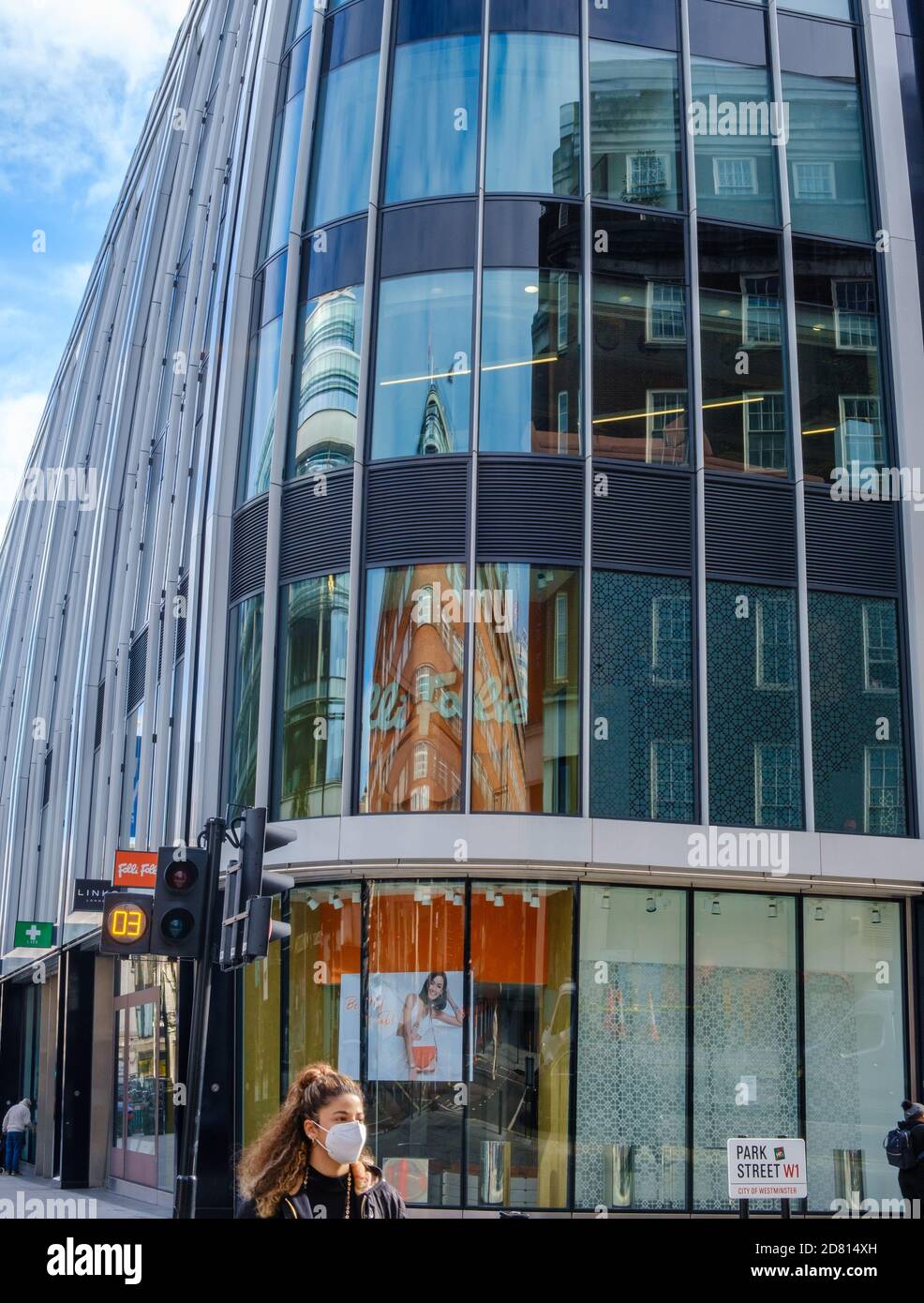 Sun reflects on glass walls of Park House modern, curved building at Park Street & Oxford Street, London, while girl with face mask crosses street. Stock Photo