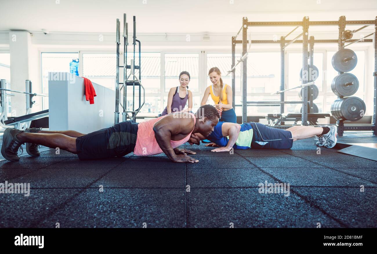 Two men have a push-up competition in the gym with girls cheering Stock Photo