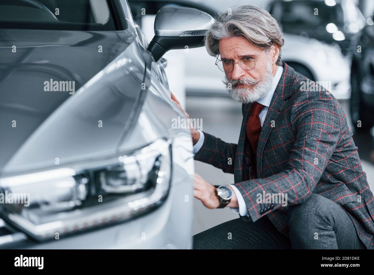 Fashionable old man with grey hair and mustache touching new car indoors in salon Stock Photo