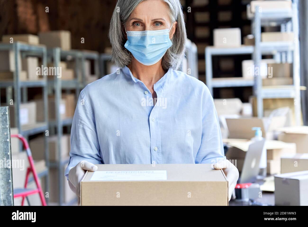 Older woman entrepreneur wearing face mask holding parcel box, portrait. Stock Photo