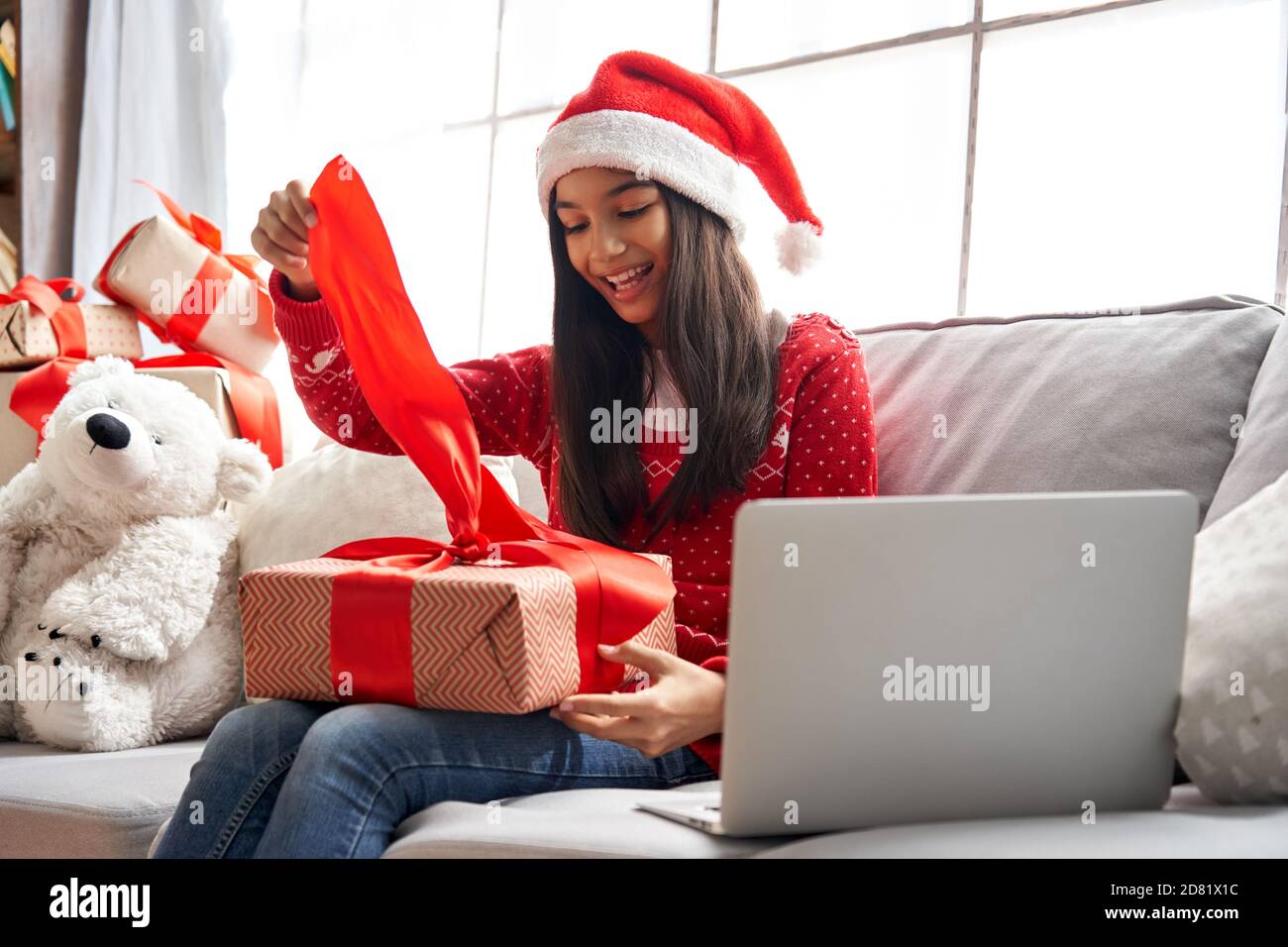 Excited indian girl wear santa hat opens gift video calling family on Christmas. Stock Photo