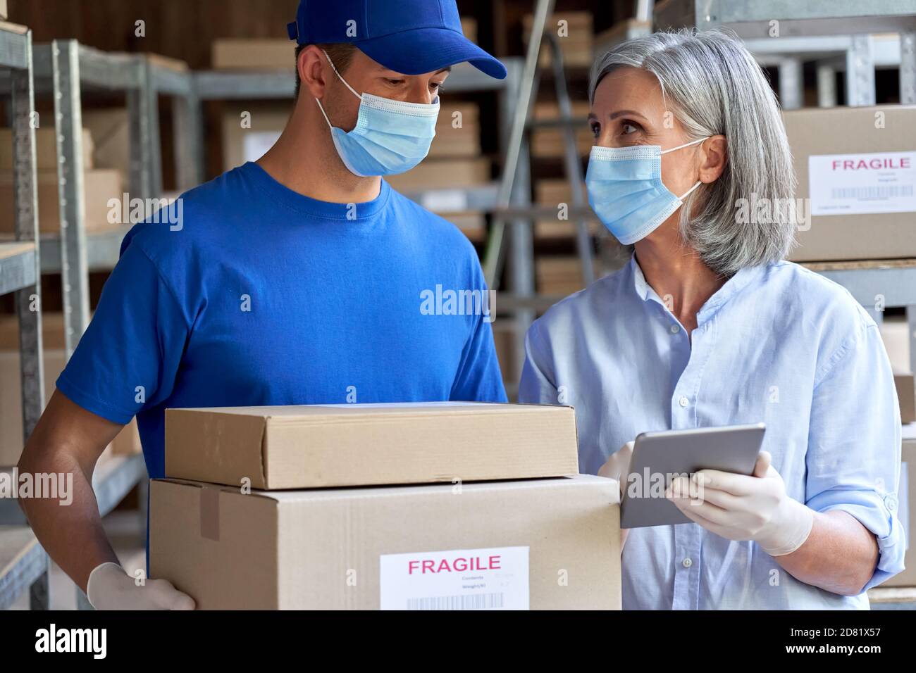 Female manager and courier wearing face masks talking in warehouse. Stock Photo