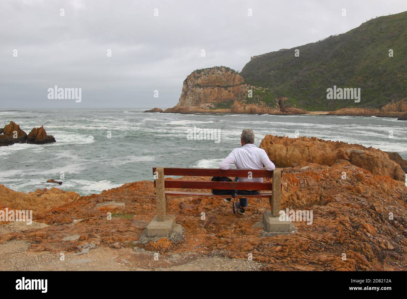 A man sits on a bench, looking at the sandstone cliffs of Knysna Heads. A famous landmark along the renowned Garden Route, South Africa, Africa. Stock Photo