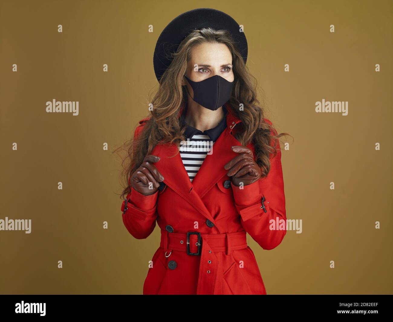 Life during coronavirus pandemic. modern woman in red coat with black mask looking into the distance against bronze background. Stock Photo