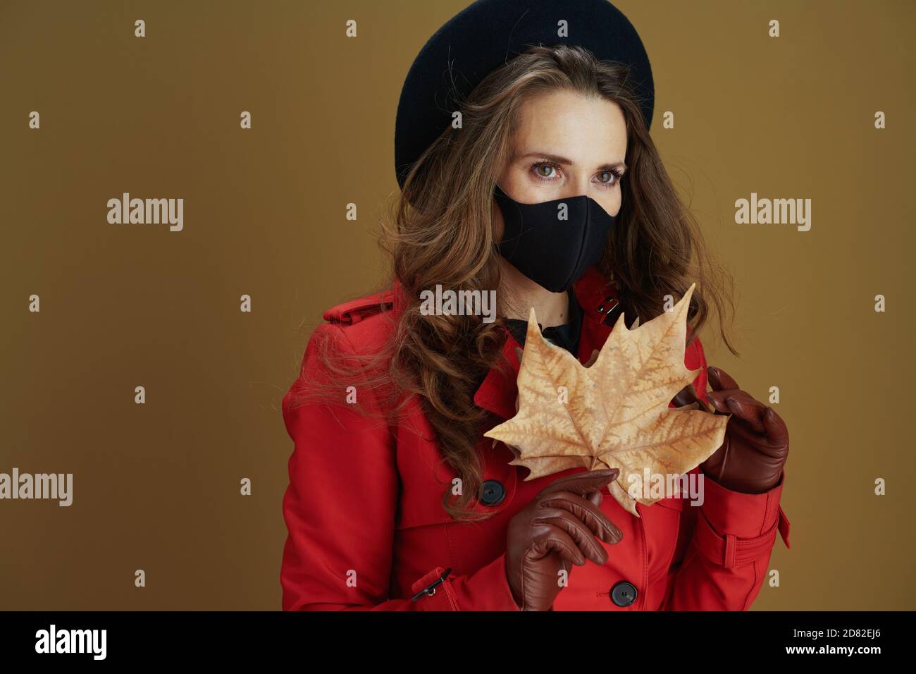 Life during covid-19 pandemic. elegant housewife in red coat with black mask and yellow autumn maple leaf isolated on beige background. Stock Photo