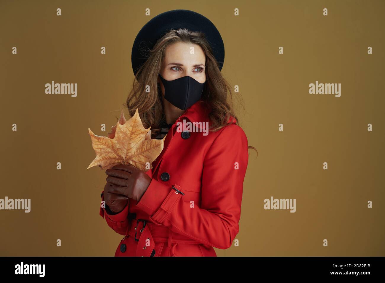 Hello autumn. trendy middle aged woman in red coat with black mask and yellow autumn maple leaf looking into the distance on beige background. Stock Photo