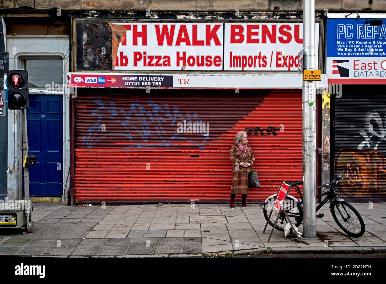 A smartly dressed mature woman waiting in front of a rundown and shuttered Pizza shop on Leith Walk, Edinburgh, Scotland, UK. Stock Photo