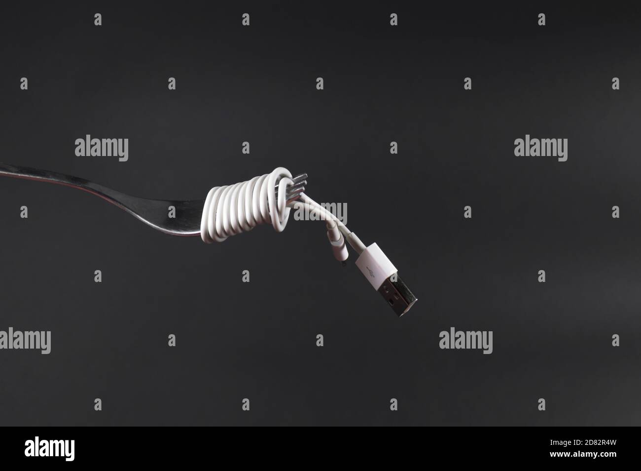Charging mobile phone cable wrapped around fork like spaghetti. Dark grey background with fork and power cable in front. Conceptual photograph. Stock Photo