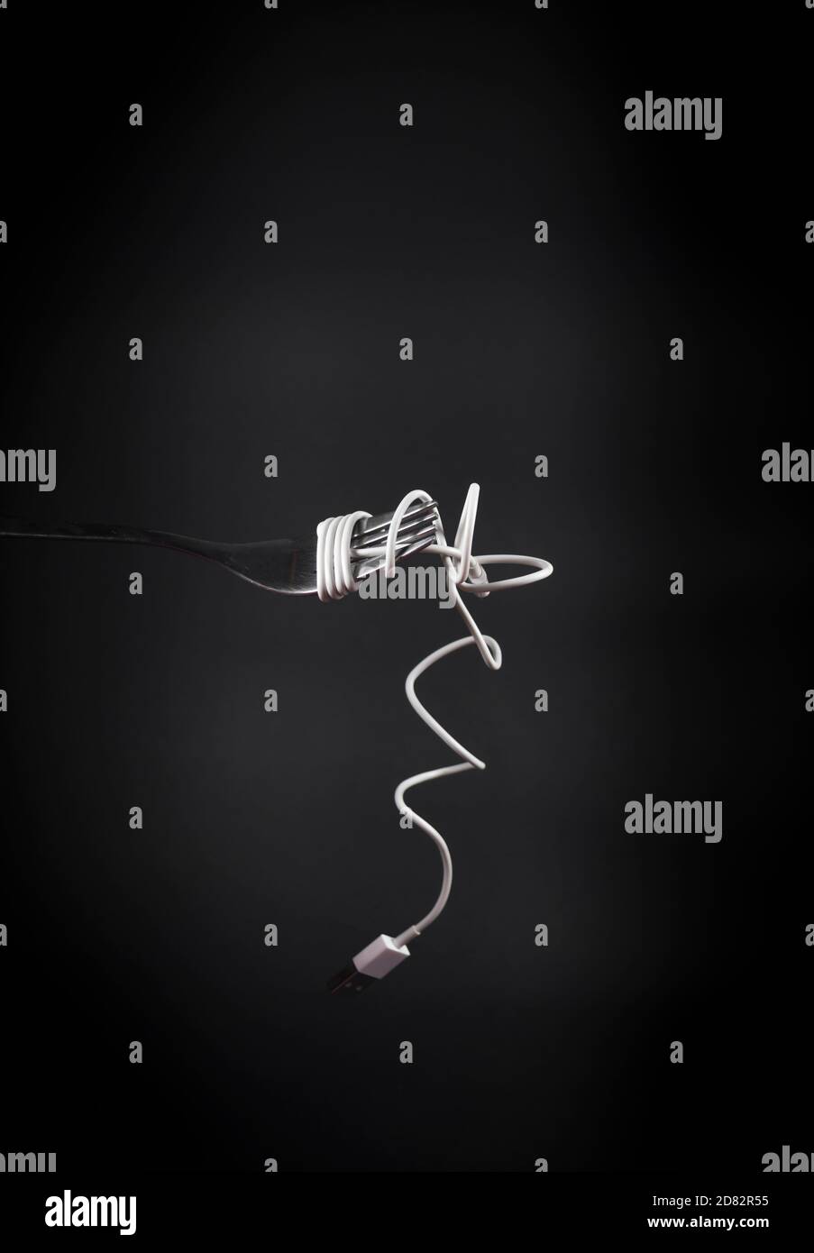 Charging mobile phone cable wrapped around fork like spaghetti. Dark grey background with fork and power cable in front. Conceptual photograph. Stock Photo