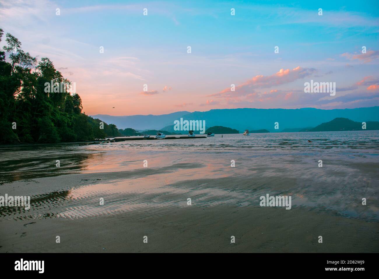 Landscape of a beach at sunset with boats on the water and some trees ...