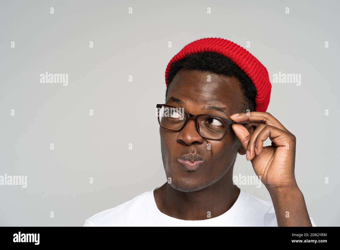 Close up studio portrait of unsure doubtful Afro American man in red hat holding eyeglasses, peers, suspicious looking away at blank copy space for ad Stock Photo