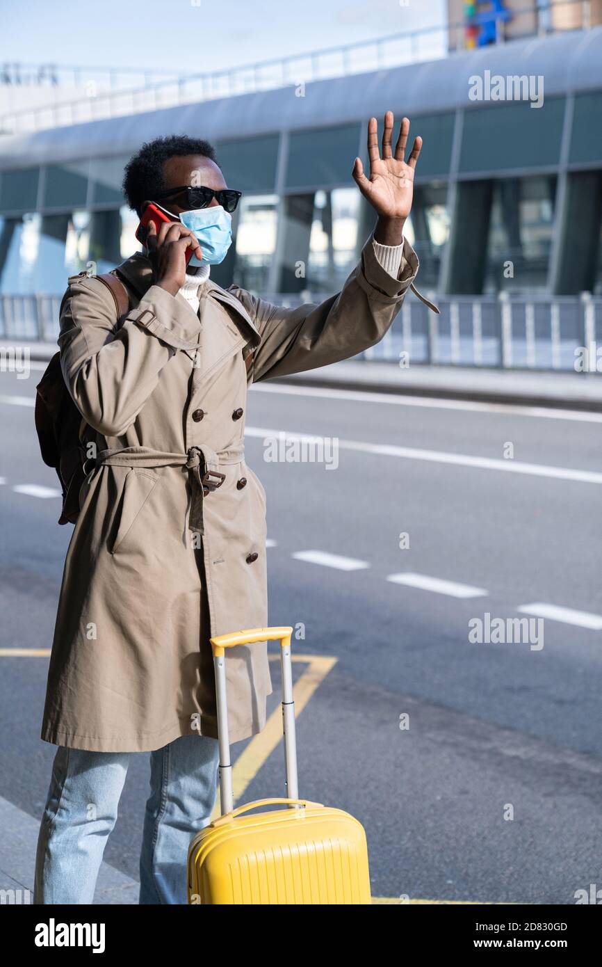 Afro-American traveler man with suitcase wear face mask stands in airport terminal, talking on mobile phone, calling taxi, raising hand. waiting for c Stock Photo