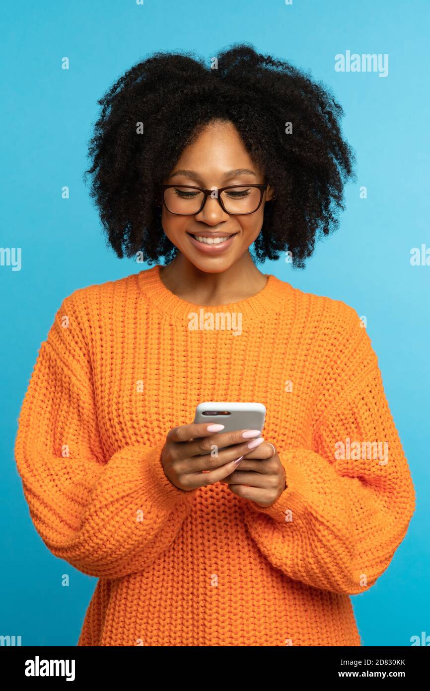 Stylish happy dark skinned millennial woman with curly hair wear orange knitted jumper, reading message from boyfriend, chatting in social media, text Stock Photo