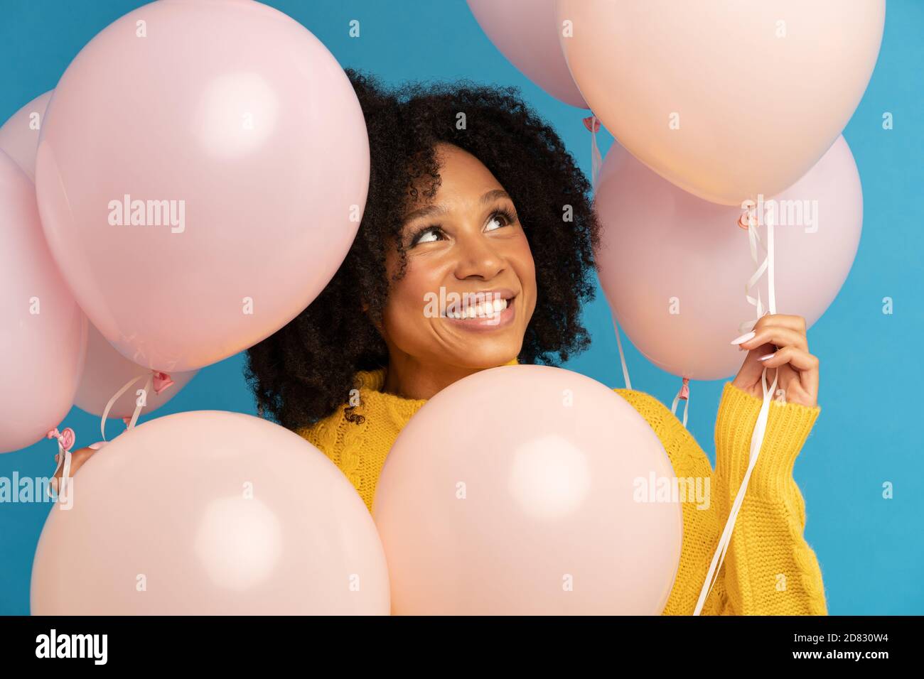 Close up of happy dark skinned woman among pastel pink air balloons with joy and happiness, celebrates birthday, looking up, wears yellow jumper, stan Stock Photo